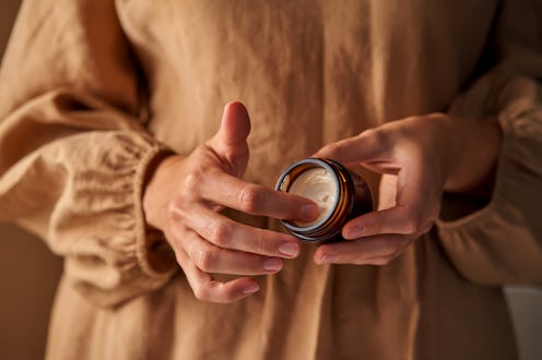 A girl in a beige dress holds an open natural cosmetic cream in her hands.