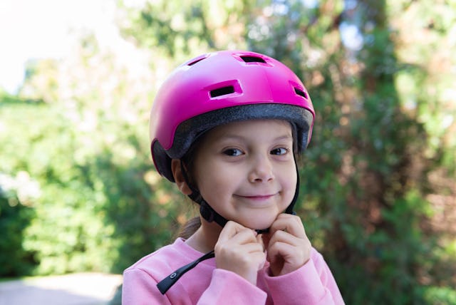 A close-up of a young girl smiling confidently while adjusting her bright pink helmet. She is outdoo...