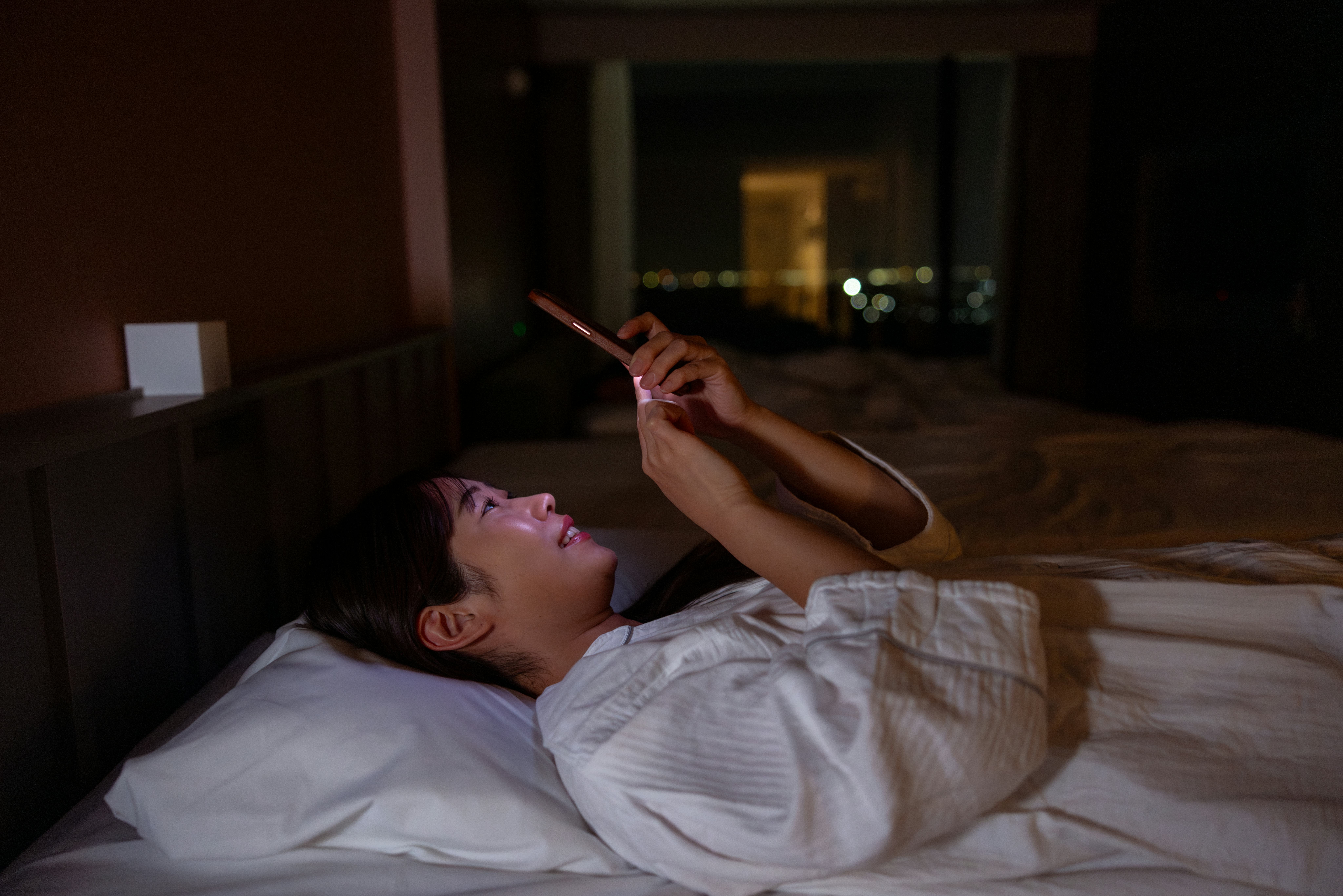 Woman in pajamas lying on back and using smart phone in hotel room at night