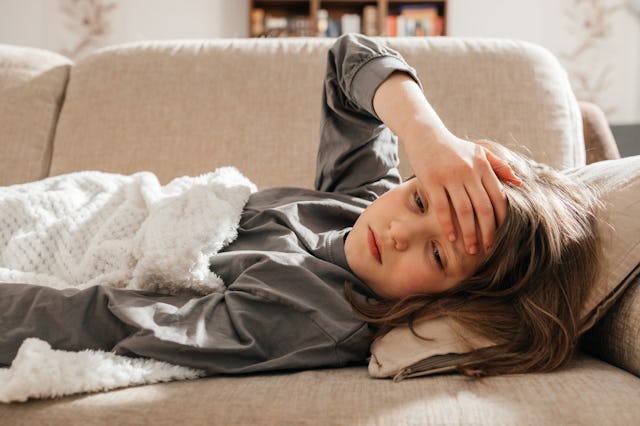 A sick dark-haired teenage girl lies on the sofa, covered with a blanket and put her hand on her for...