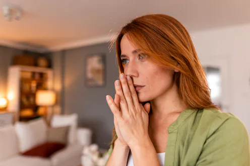 Thoughtful redhead woman with hands clasped, gazing away while standing in a softly blurred living r...