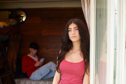 She is leaning in the doorway of an eco lodge with her boyfriend sitting inside using his smart phon...