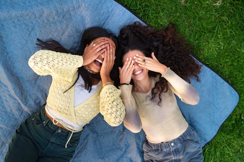 Two joyful young women relaxing on a blanket in a park, playfully hiding their faces with their hand...
