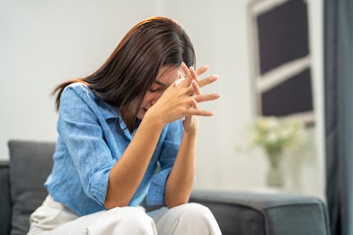 A young woman in casual clothing sits at home, head resting on hands, reflecting deeply.