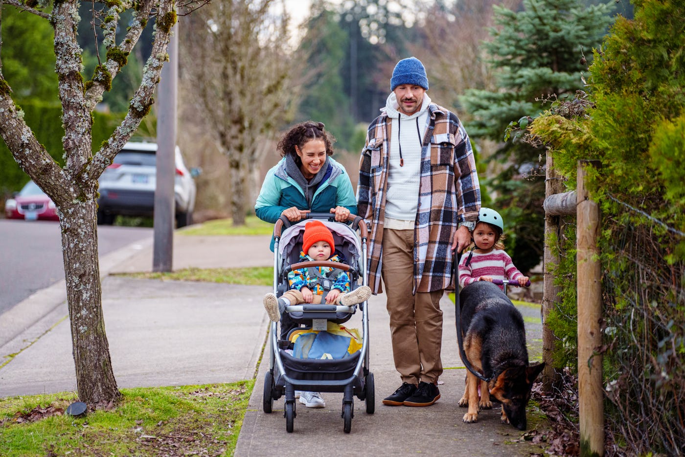 A cheerful Eurasian woman pushes her toddler son in a stroller while on a relaxing walk with her husband, four year old daughter and German Shepard pet dog along a sidewalk in their residential neighborhood located in Oregon on a cool, winter day.