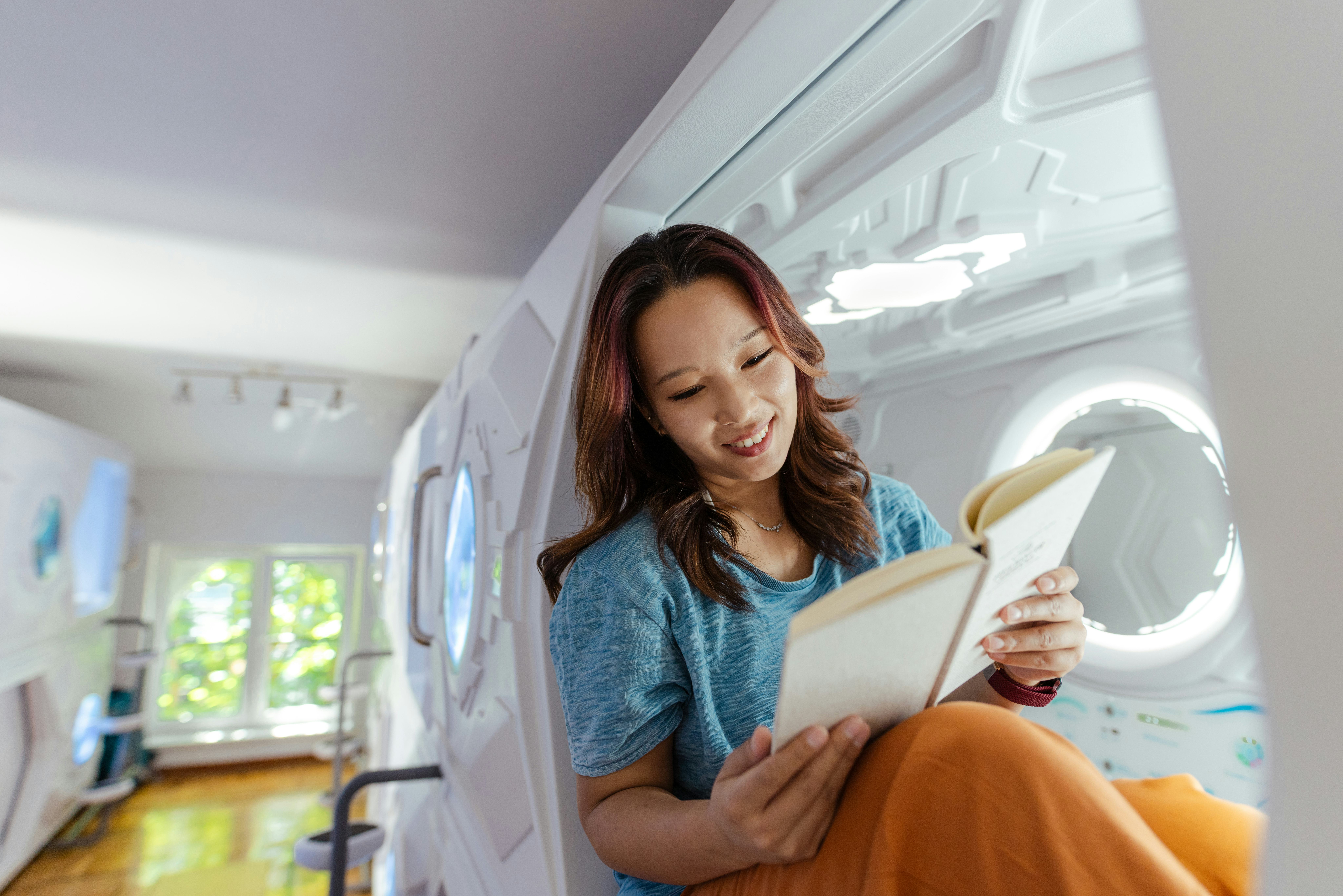 A smiling young woman relaxes and reads a book in a modern capsule-like hostel room, showcasing a un...