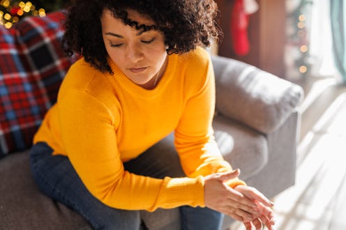 Portrait of worried young woman sitting on the sofa, rubbing her hands together, anxiously contempla...