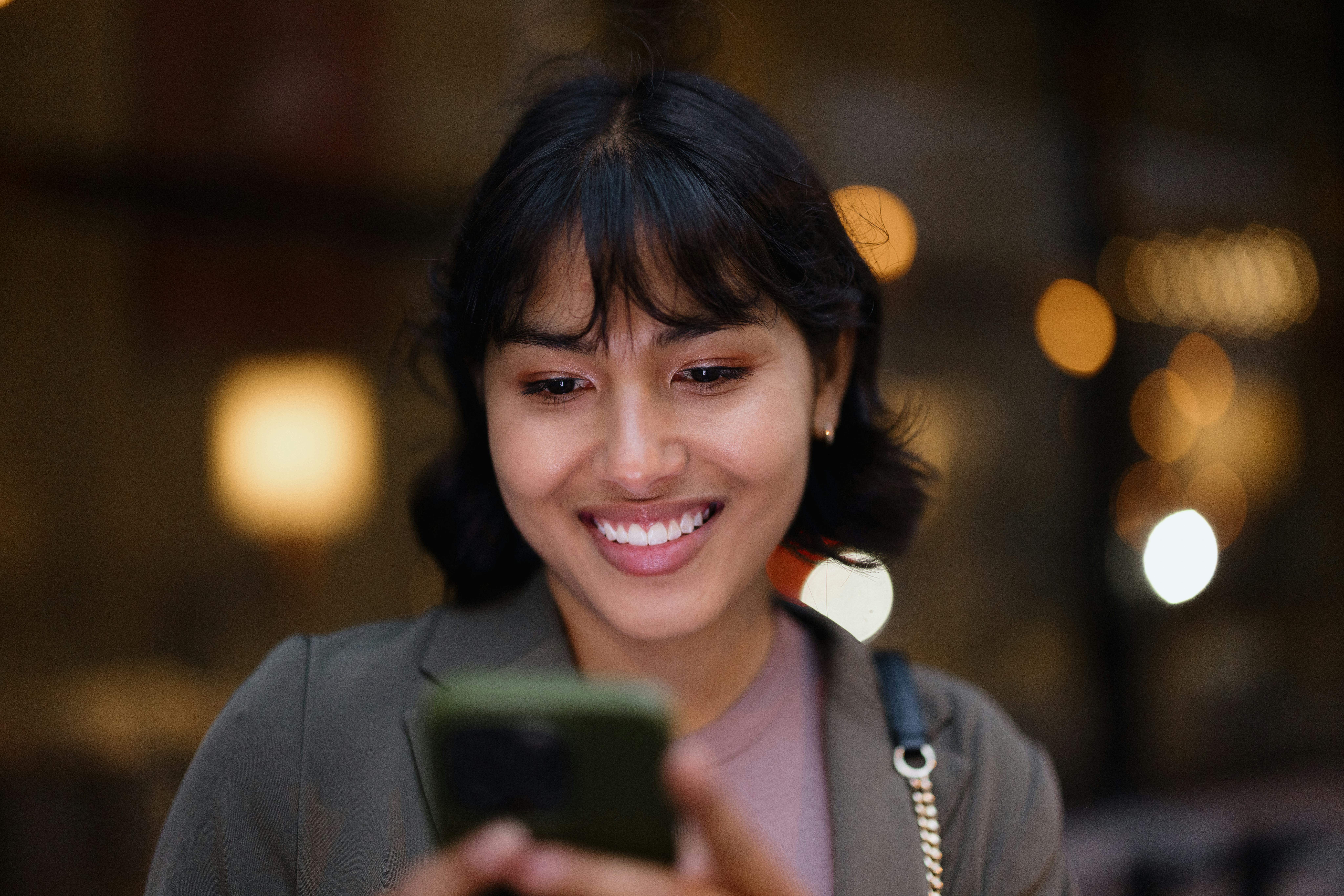 Portrait of a cheerful young businesswoman smiling while using a mobile phone in the vibrant city at...