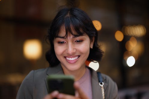 Portrait of a cheerful young businesswoman smiling while using a mobile phone in the vibrant city at...