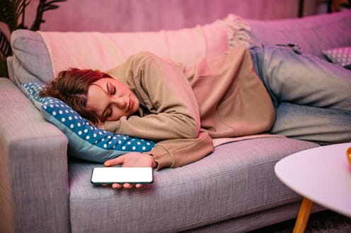 Young woman falling asleep while using her phone on the sofa at night.