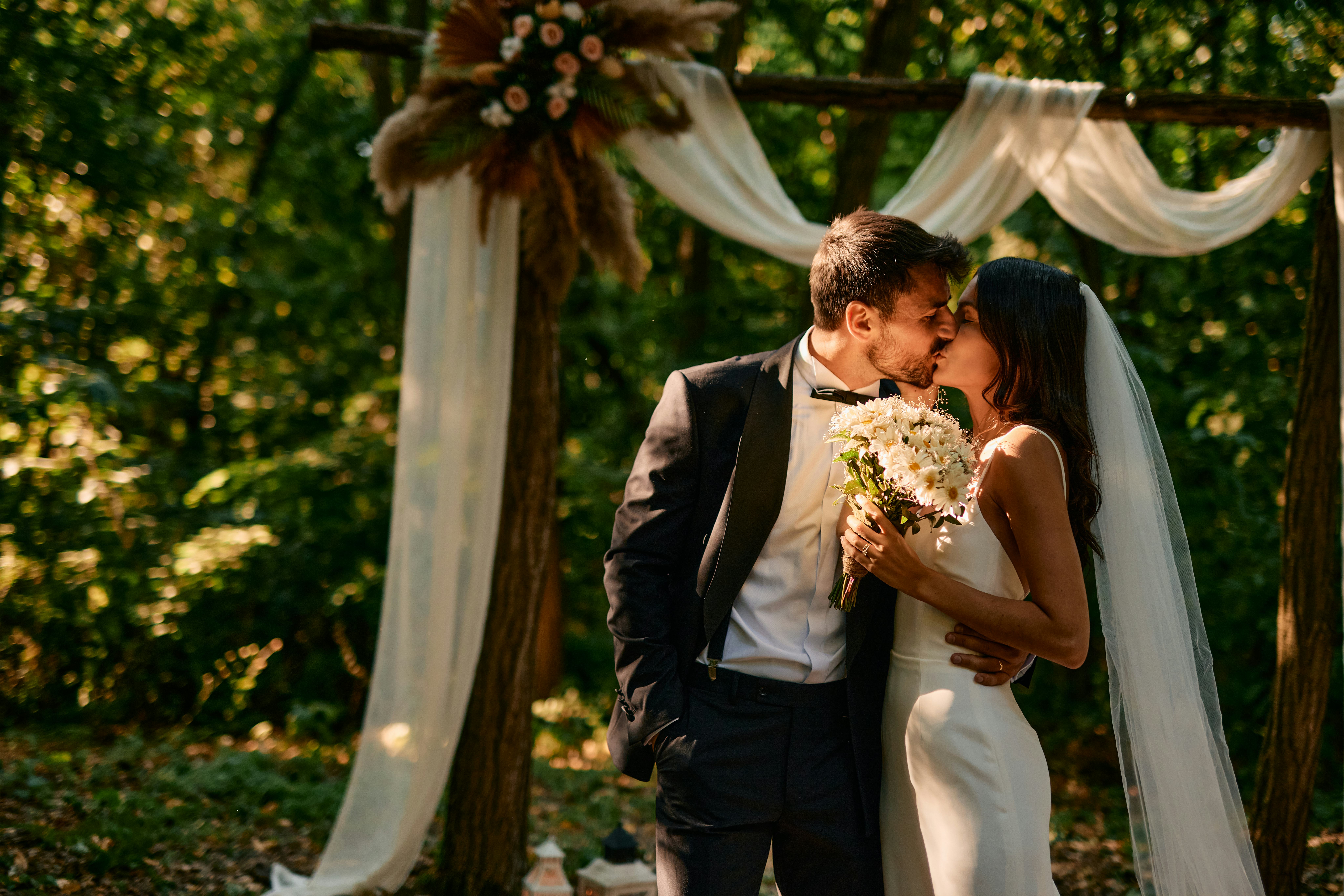 Bride and groom are kissing during their romantic outdoor wedding ceremony in a beautiful forest set...