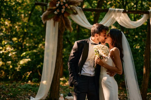 Bride and groom are kissing during their romantic outdoor wedding ceremony in a beautiful forest set...