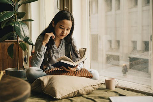 A warm, peaceful scene of a woman reading a book beside a sunny window, surrounded by cushions and a...
