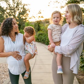 Two young mothers talk happily as they hold their toddler daughters on their hips and walk through t...