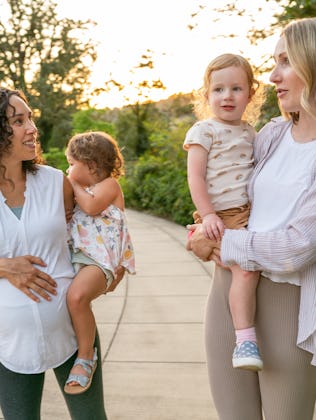 Two young mothers talk happily as they hold their toddler daughters on their hips and walk through t...