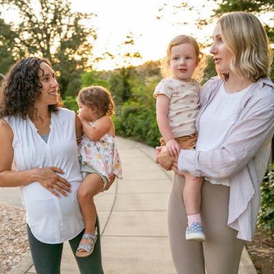 Two young mothers talk happily as they hold their toddler daughters on their hips and walk through the park at sunset. One of the women is eight months pregnant.