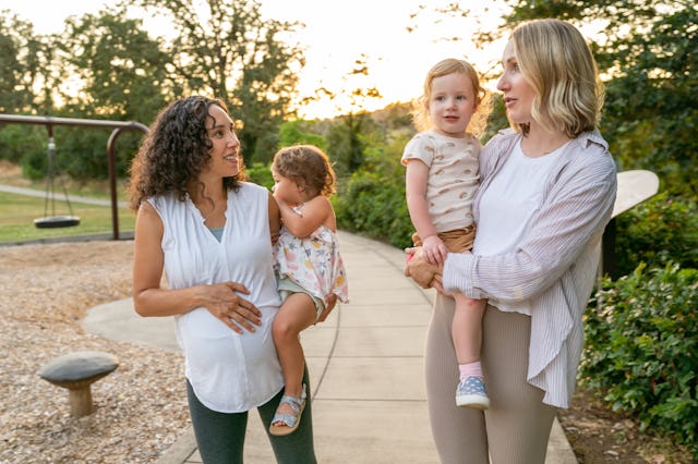 Two young mothers talk happily as they hold their toddler daughters on their hips and walk through t...