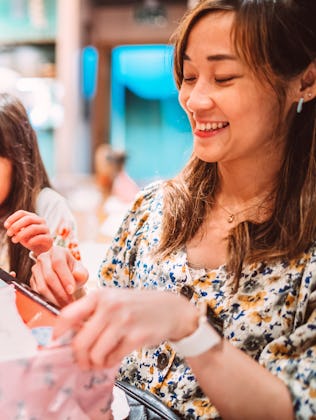 A teenage girl unwraps her birthday present with wide-eyed excitement, surrounded by her smiling sis...