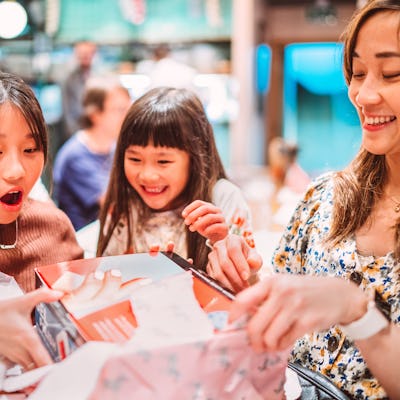 A teenage girl unwraps her birthday present with wide-eyed excitement, surrounded by her smiling sister and mother.