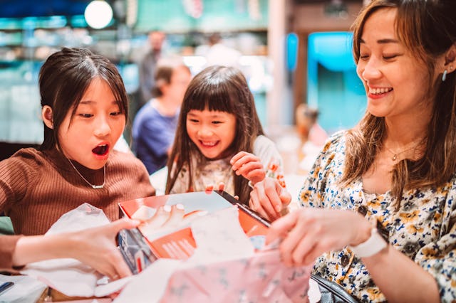 A teenage girl unwraps her birthday present with wide-eyed excitement, surrounded by her smiling sis...