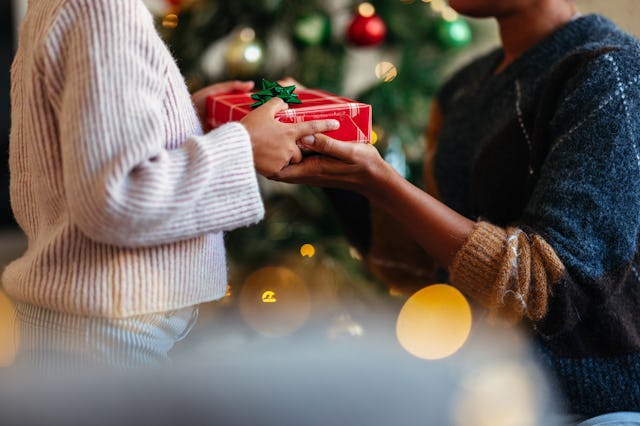A close-up of hands exchanging a wrapped Christmas gift, set in front of a decorated tree. The cozy ...