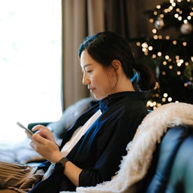 Smiling young Asian woman sitting on sofa in the living room next to a decorated Christmas tree and ...