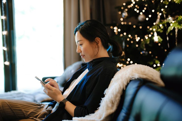 Smiling young Asian woman sitting on sofa in the living room next to a decorated Christmas tree and ...