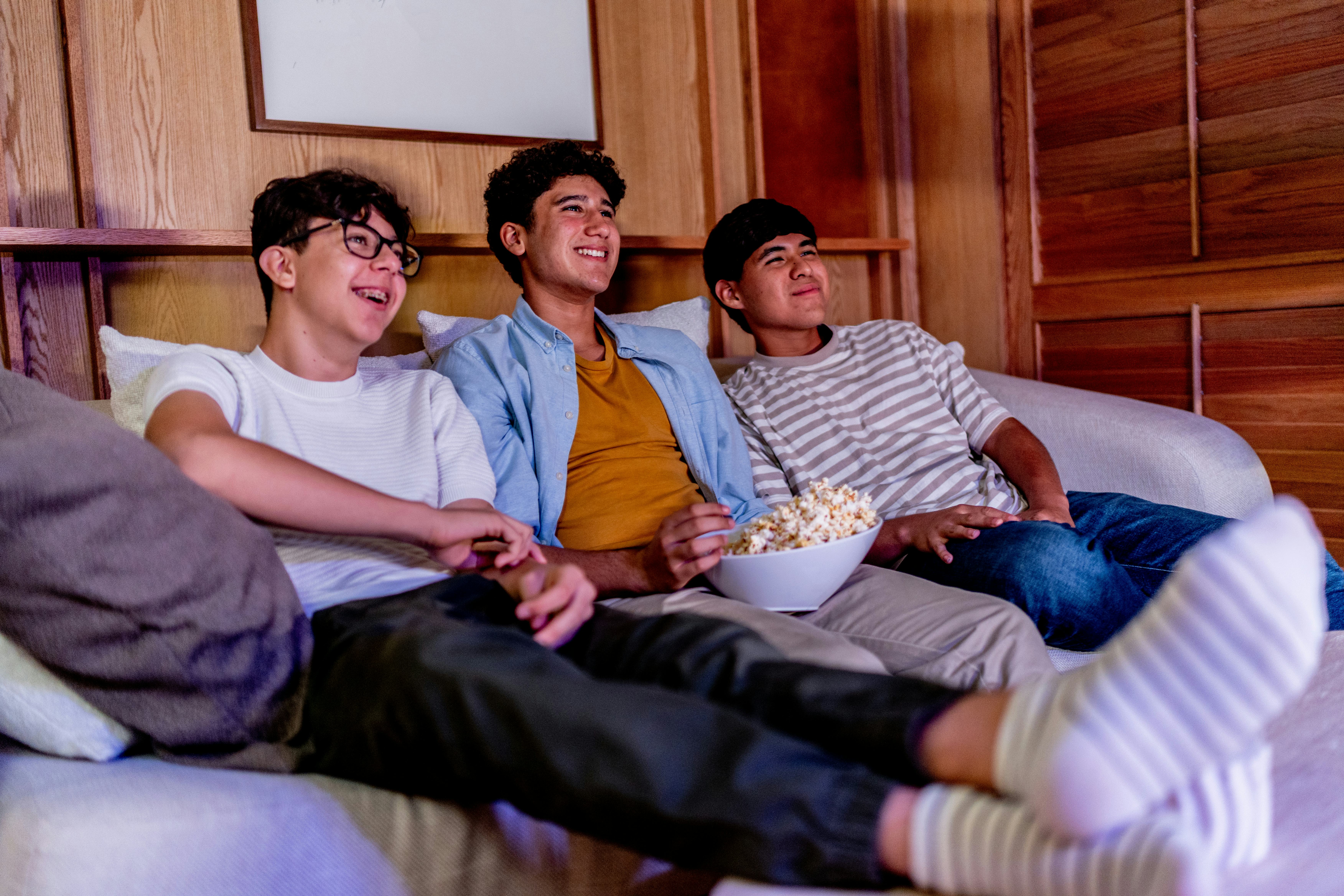 Teenage boys talking and eating popcorn while watching movie at home.