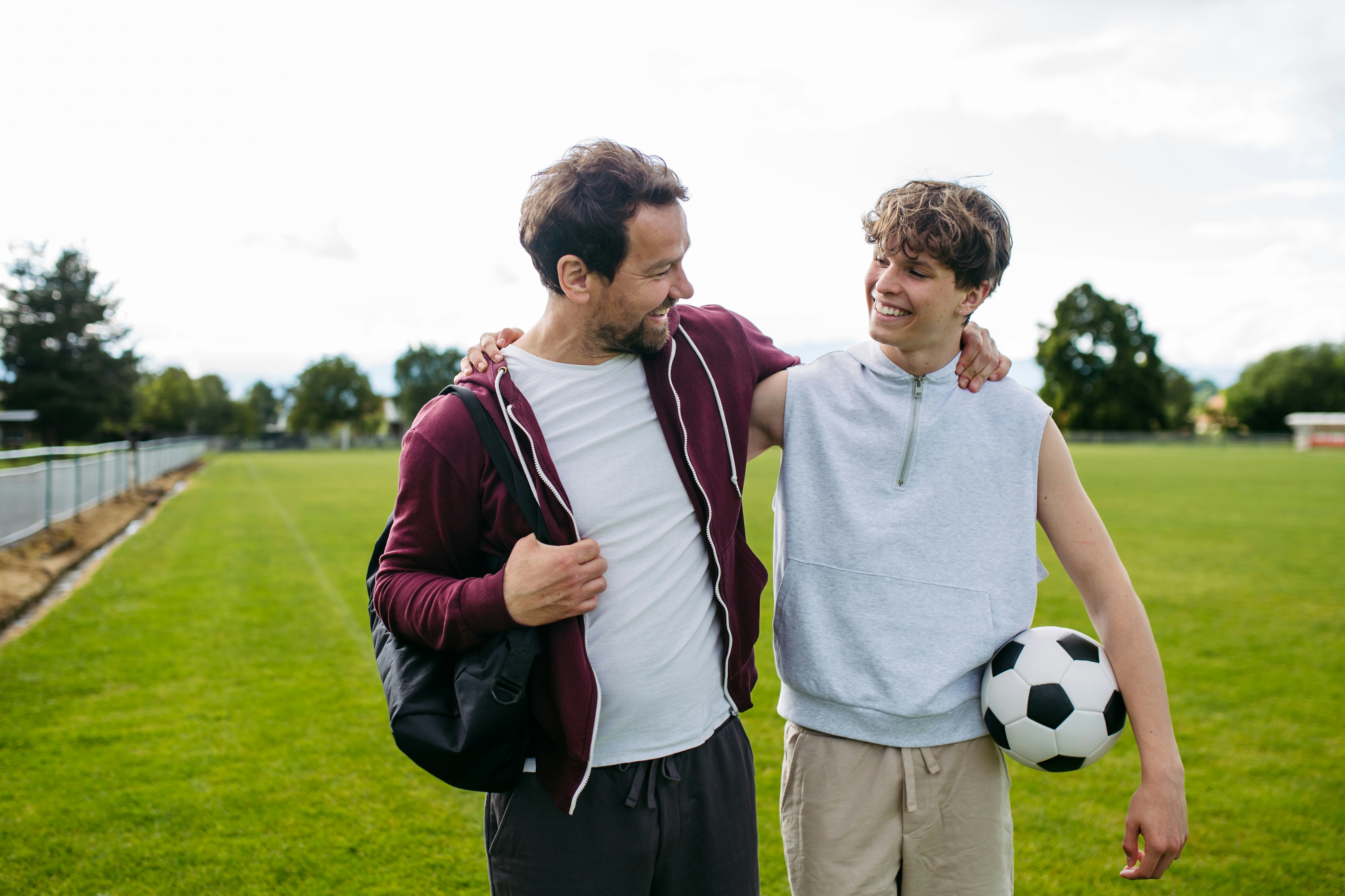 Father and teenage son playing football on a field at sunset.