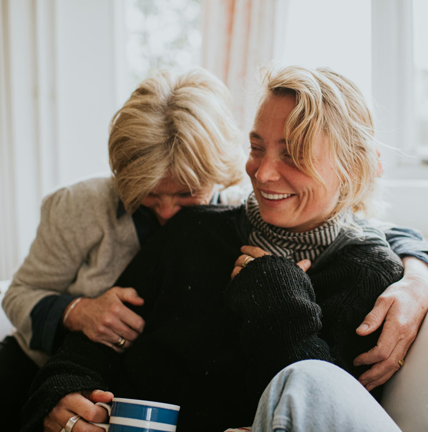 A mother hugs her adult daughter in a domestic living room. The scene is comfortable and loving as the older woman embraces the woman in a loving hug. Represents human relationships.