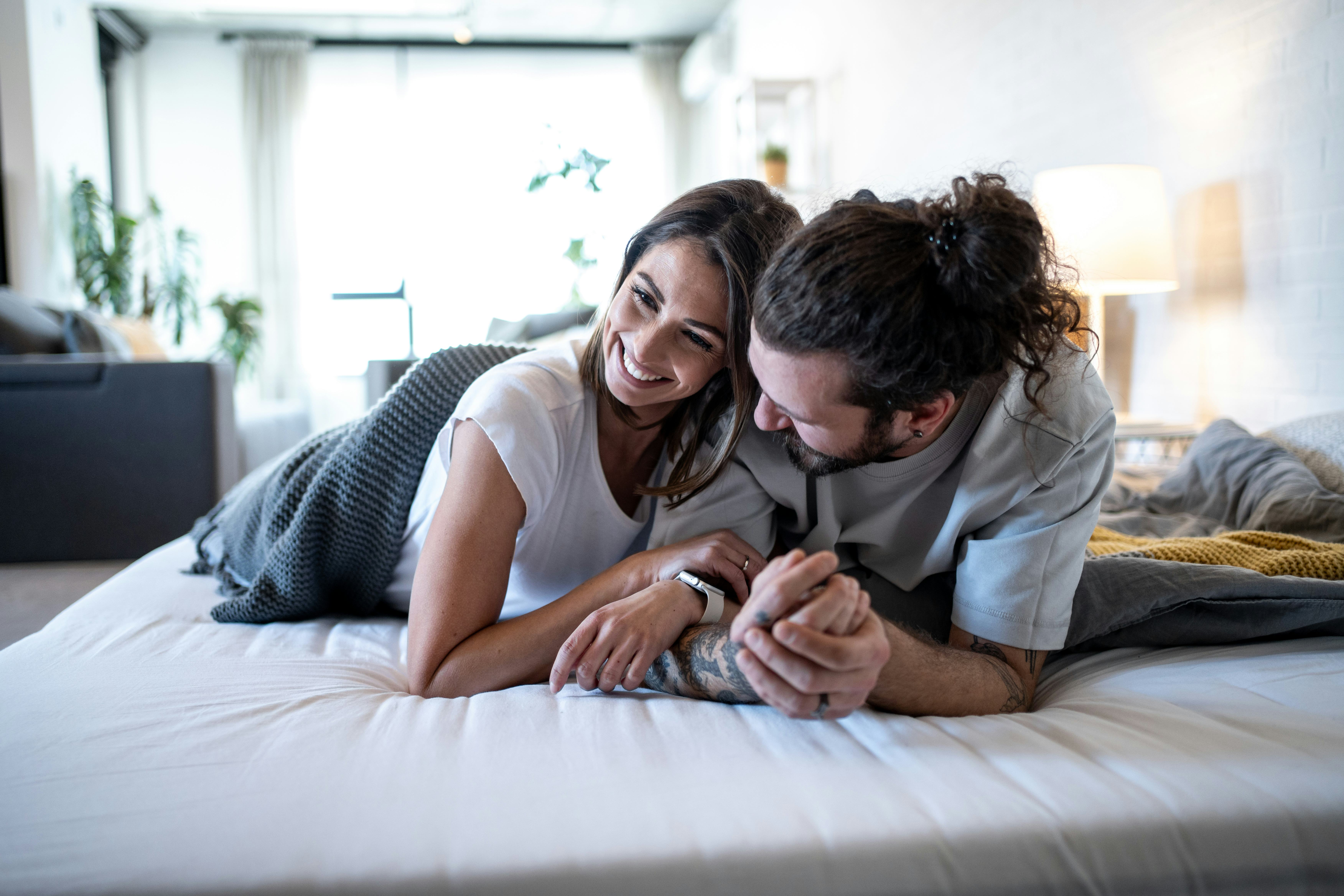 Young couple lying on a cozy bed, holding hands and sharing smiles while gazing into each other's ey...