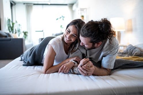 Young couple lying on a cozy bed, holding hands and sharing smiles while gazing into each other's ey...