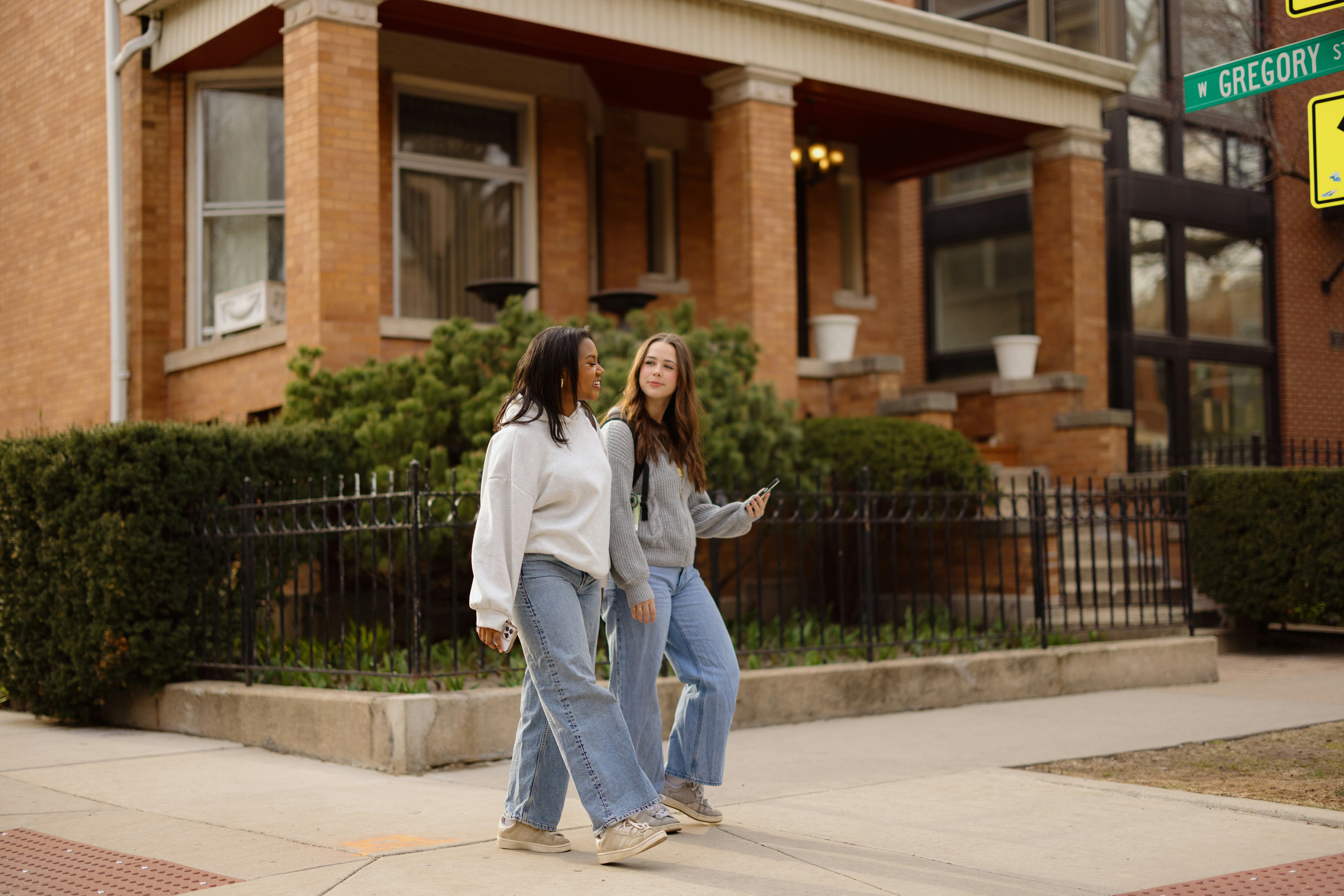 Two female students walking on the sidewalk in front of a brick house, enjoying a friendly conversat...