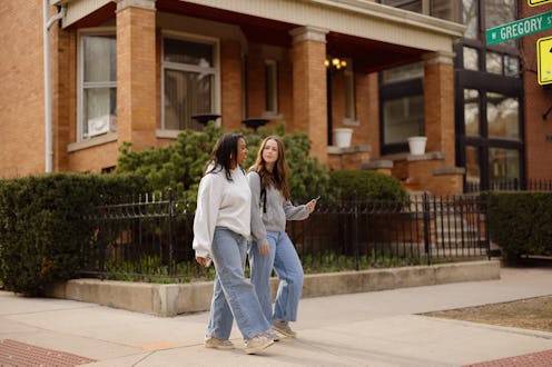 Two female students walking on the sidewalk in front of a brick house, enjoying a friendly conversat...