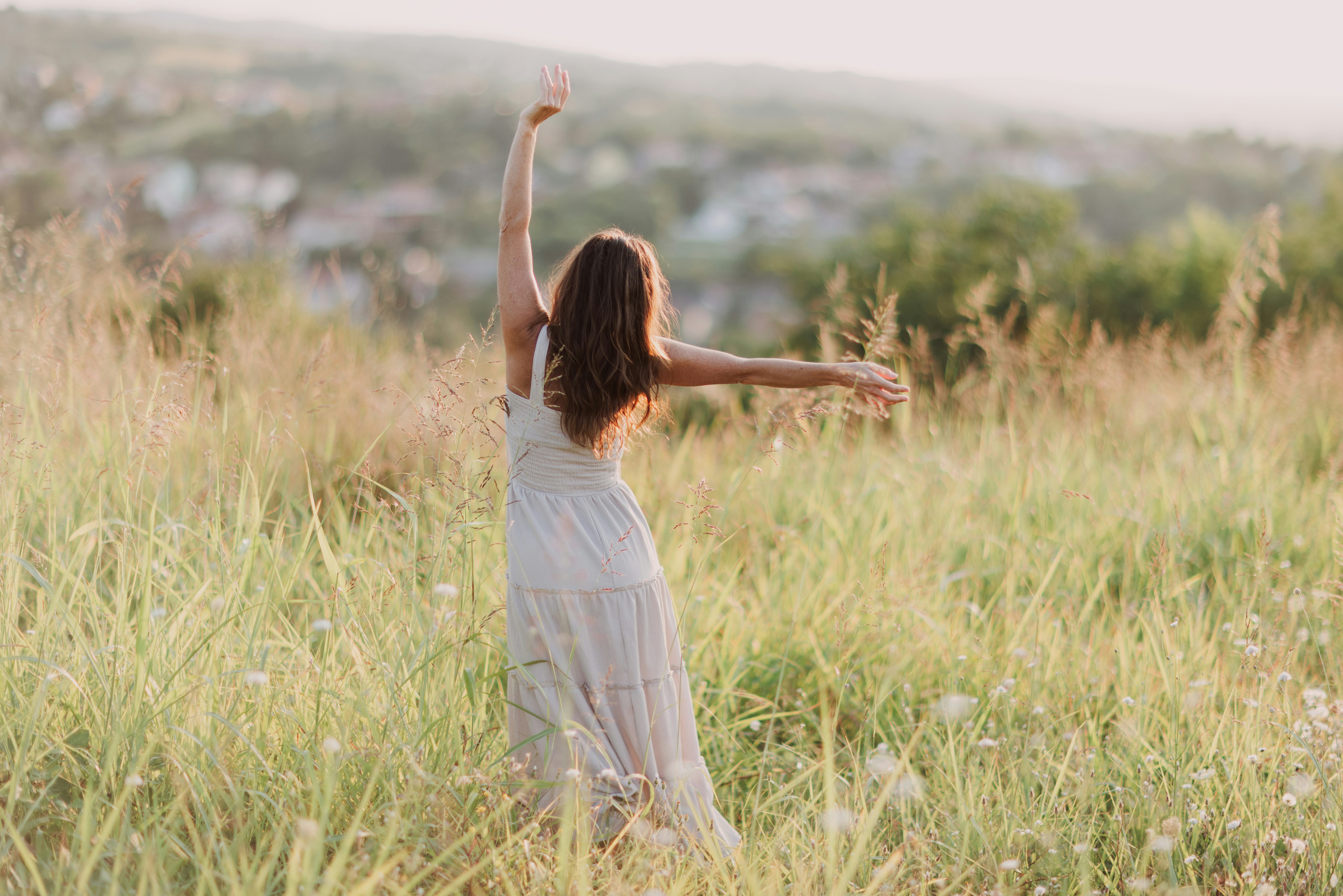 A young woman joyfully dances in a green meadow under soft sunlight, surrounded by nature.