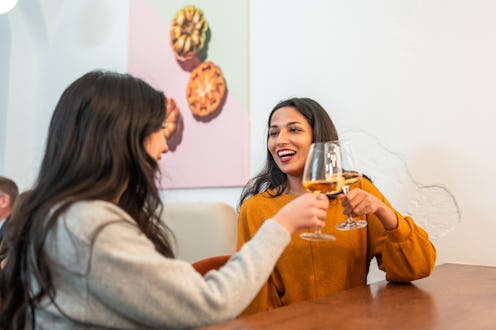 Candid portrait of young Indian and Hispanic women friends toasting wine glasses, sharing genuine la...