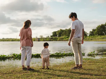 A mother and father stand along a riverbank with their child.