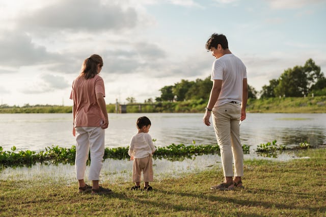 A mother and father stand along a riverbank with their child.