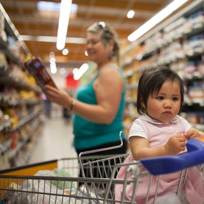 A baby sits in a shopping cart at the supermarket while her family members looks at food prices.