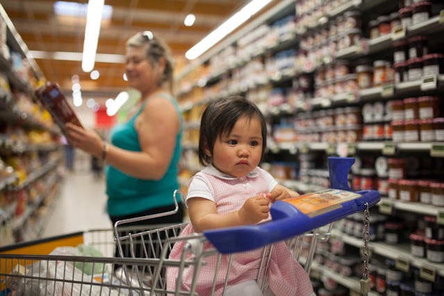 A baby sits in a shopping cart at the supermarket while her family members looks at food prices.