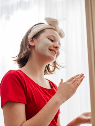 Teenage girl takes care of her face skin at home, applies a cosmetic clay mask to her face. Self-car...