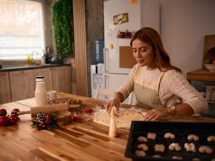 Young woman baking Christmas cookies in a cozy kitchen, using festive cookie cutters and dough, surr...