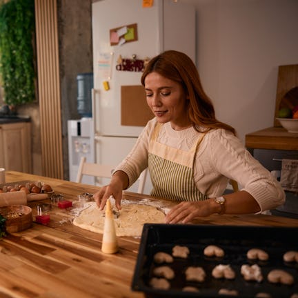 Young woman baking Christmas cookies in a cozy kitchen, using festive cookie cutters and dough, surr...