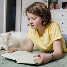 Young girl reading a book with her white dog sleeping nearby in her bedroom