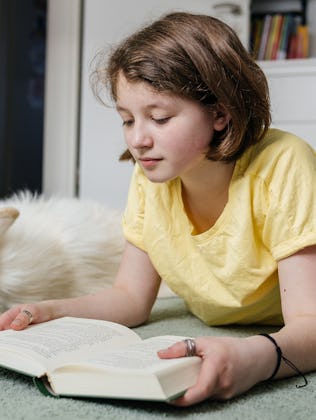 Young girl reading a book with her white dog sleeping nearby in her bedroom