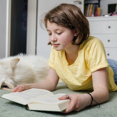Young girl reading a book with her white dog sleeping nearby in her bedroom