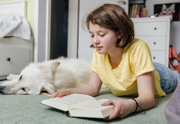 Young girl reading a book with her white dog sleeping nearby in her bedroom