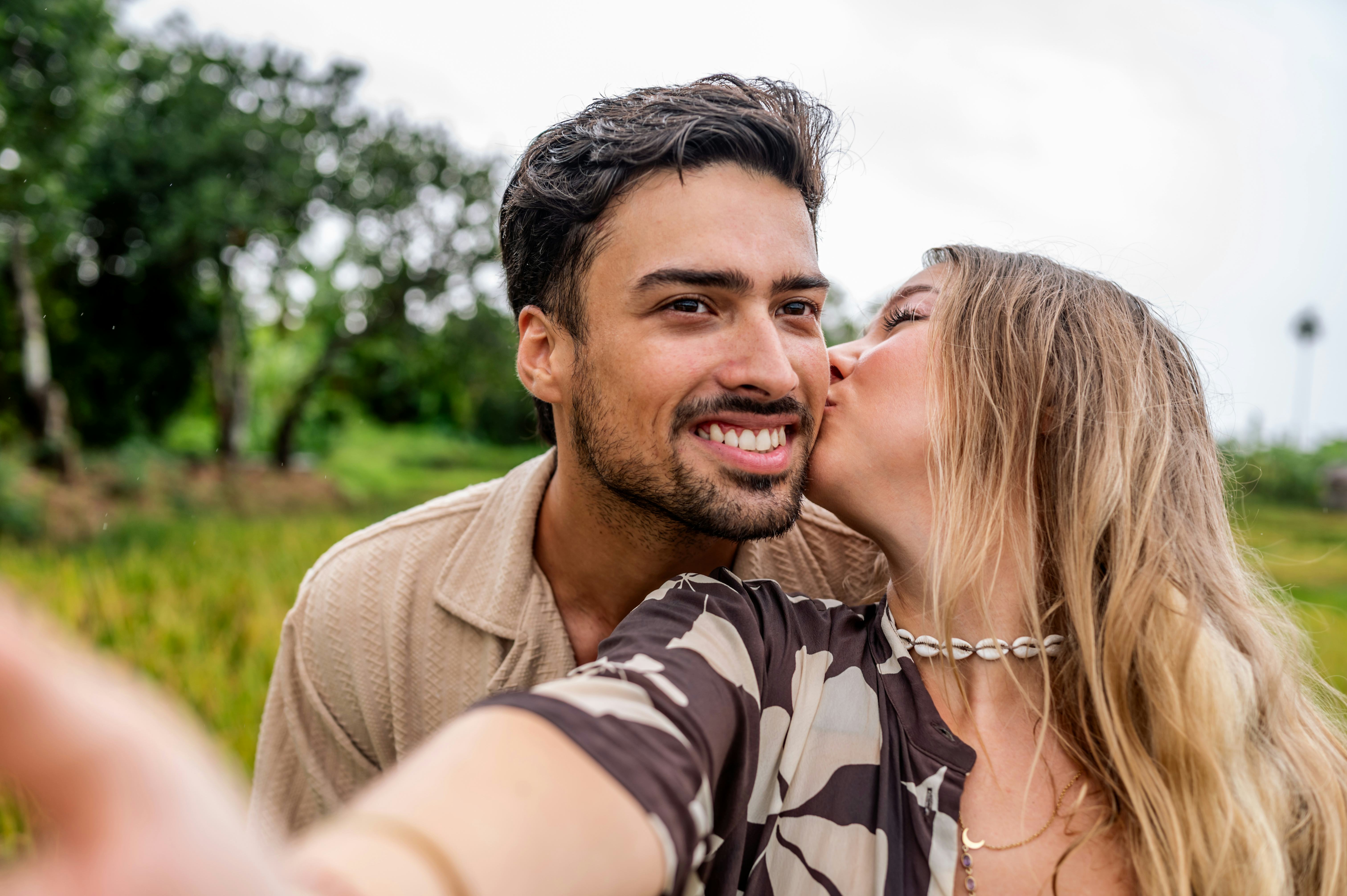 A smiling man and woman share a kiss in a sunny outdoor setting, capturing affection and connection....