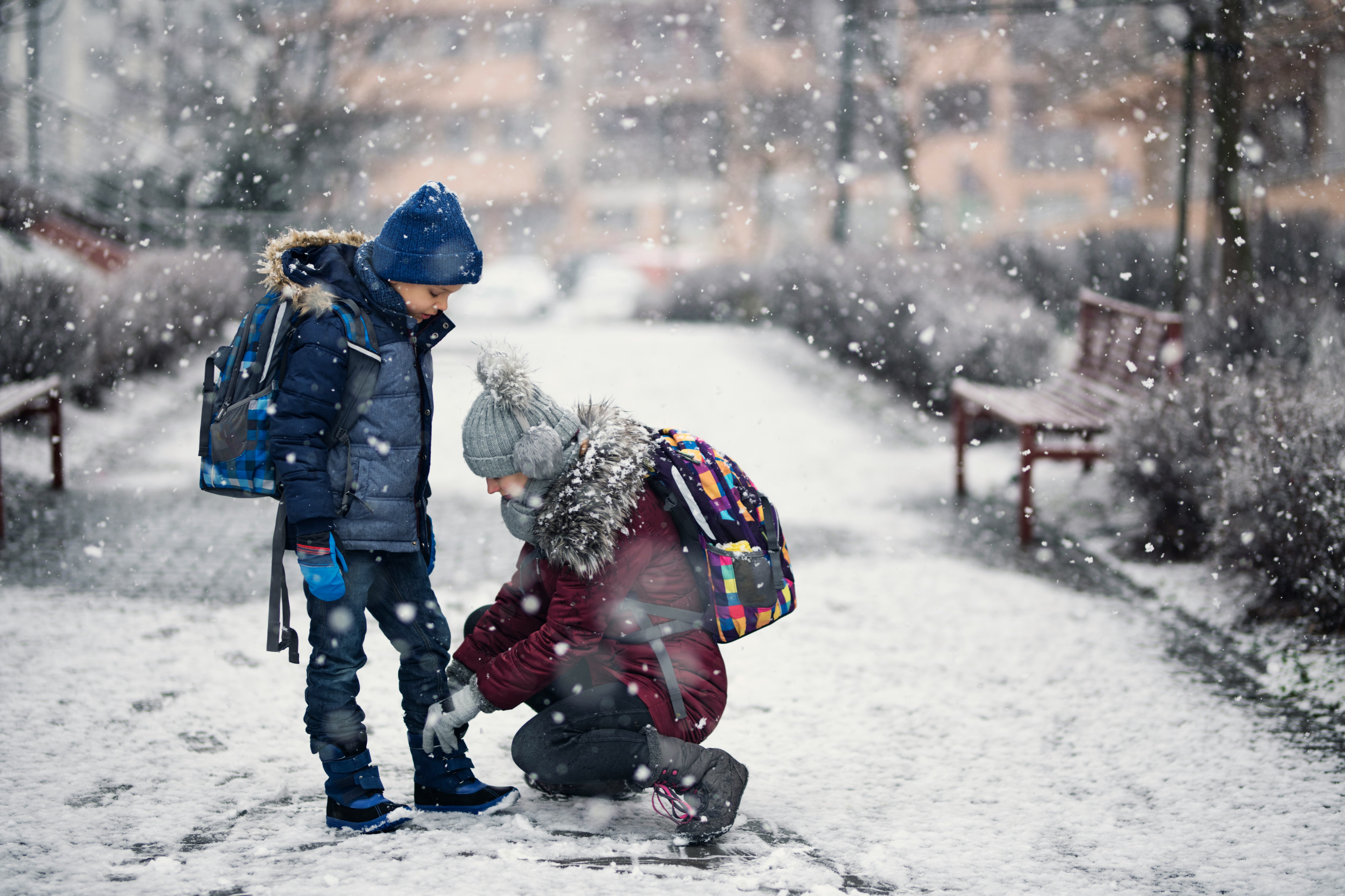 A girl stops to tie her brother's shoe as they walk to school in the snow. 