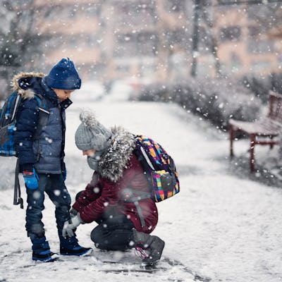 A girl stops to tie her brother's shoe as they walk to school in the snow.